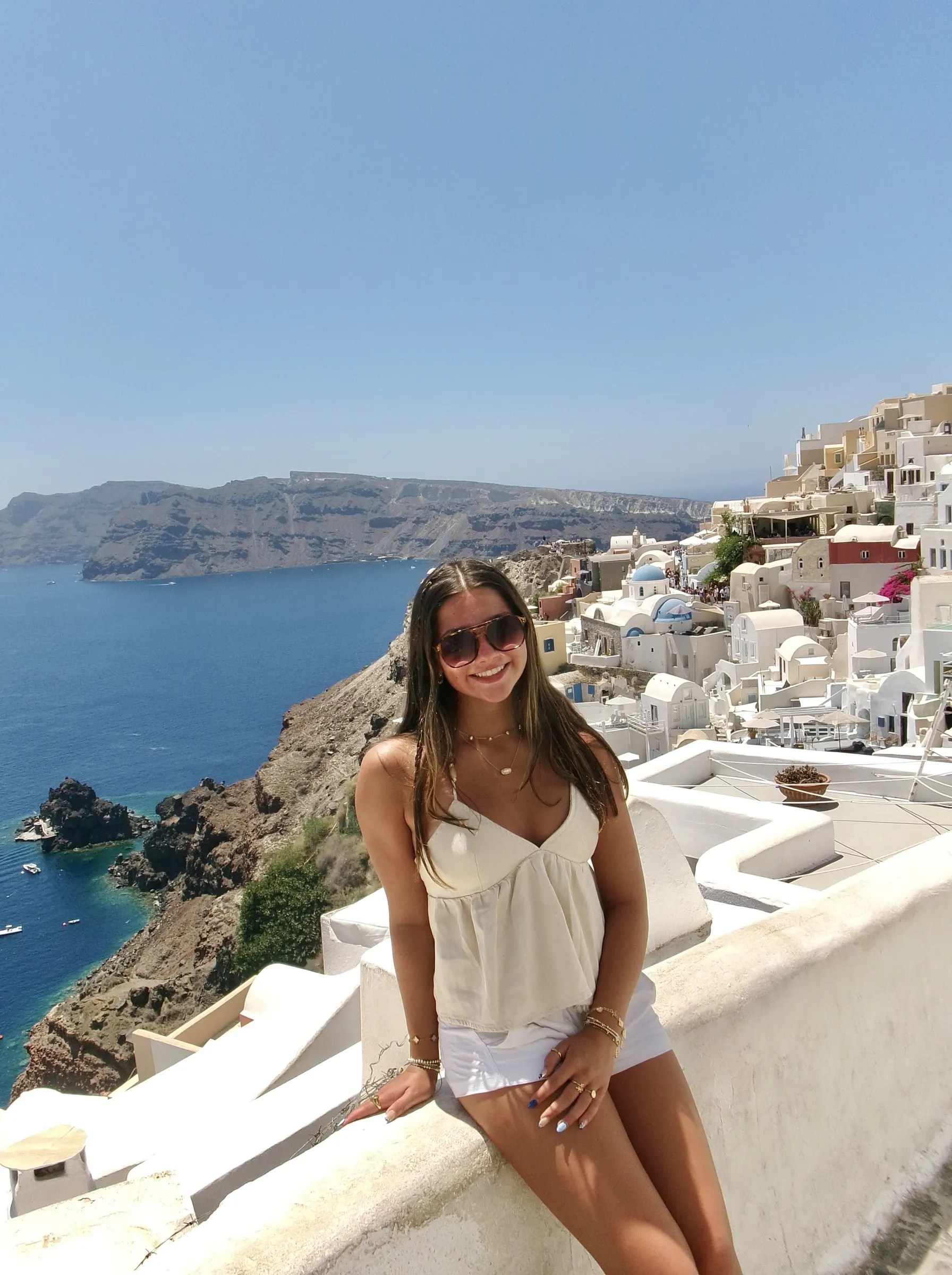 Original photo: a smiling woman in sunglasses sits on a white wall above the sea in Santorini on a clear day